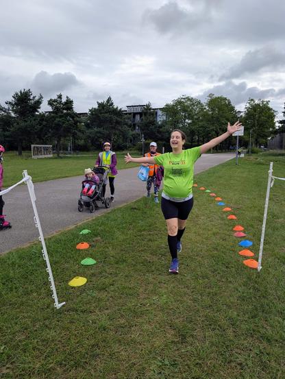 A woman in a green shirt crosses the finish line at parkrun, arms raised in celebration. Behind her, other participants and a person pushing a stroller are visible. The finish line is marked by white poles and colorful cones on the grass.