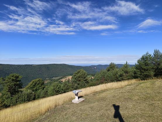 2 photos numériques couleur en format paysage, avec une vue panoramique prise depuis le socle d'une croix dont on voit l'ombre sur l'herbe. On voit la table d'orientation, de l'herbe jaune, des arbres et au loin des reliefs boisés avec une falaise. Le ciel est bleu avec une bande de nuages près des reliefs et un nuage blanc dispersé en altitude.