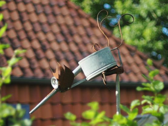 Ein Kunstwerk aus Metall ist auf einem langen Stab angebracht. Es ist aus Metall, der Körper besteht aus einem alten Topf oder AO etwas, dee Kopf, die Beine und die Schwanzfedern sind verrostet. Im Vordergrund sind unscharf Äste mit grünen Blättern am Rand zu sehen, im Hintergrund das rote Dach eines Hauses.