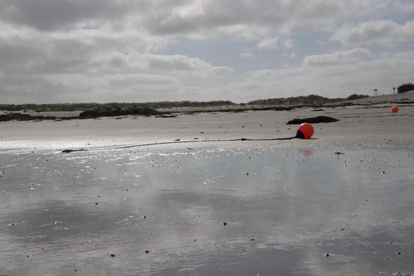Am Strand unter einem bewölkten Himmel, aber im Sonnenschein, liegt eine Boje. Das Tau an dem sie befestigt ist zeigt an, daß der Strand bei Flut hier unter Wasser steht.