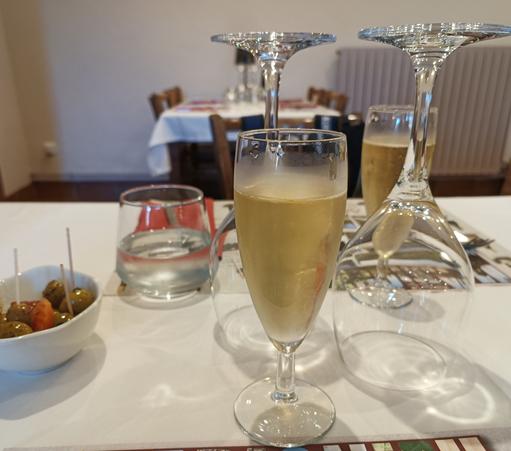 Vue rapprochée d'une table dressée avec 2 flûtes de vin blanc pétillant, des verres à eau et des verres à vin retournés sur la nappe. A gauche, un ramequin avec des olives.