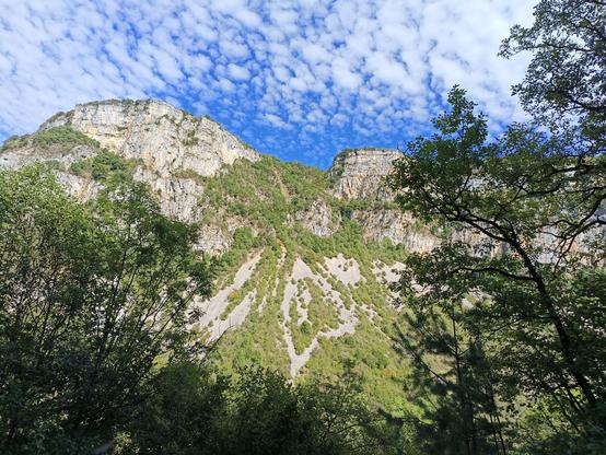 Vue prise depuis le chemin des falaises de l'autre côté de la vallée (le Crêt de la Rivoire), avec des coulées de pierres entre 2 sommets et de la végétation. Le ciel est bleu avec des nuages blancs qui moutonnent.