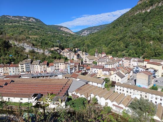 Vue prise depuis un promontoire en face de la ville de Tenay, avec une grande usine au premier plan, un habitat serré d'immeubles souvent hauts et étroits, une église, le tout dans un paysage de montagnes boisées et escarpées.