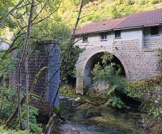 Dans le lit d'une rivière, un pilier de pont en pierre maçonnée de couleur grise. Sur la rive en face, un bâtiment avec une grande arche sur la partie inférieure, de petites fenêtres et un toit en tuiles rouges. De la végétation de la part et d'autre de la rivière.