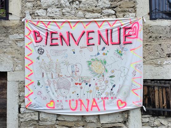 Sur le mur en pierre d'une maison à un étage, un drap blanc est accroché et décoré. En grand et en majuscules roses : bienvenue à Unaï. Il y des chevrons de couleur, des coeurs, des textes et des dessins.