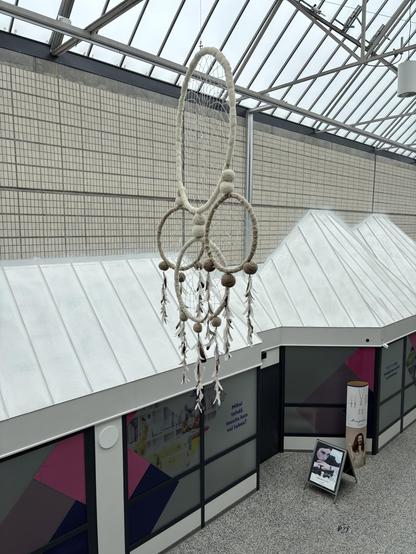 A large, intricately designed hanging mobile made of woven materials, featuring circular shapes and decorative elements like pom-poms and feathers. It is suspended from a transparent roof in an indoor space, with store windows visible below.