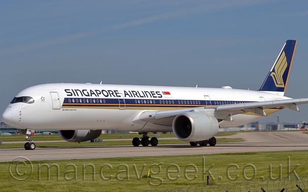 Side view of a twin engined jet airliner taxiing from right to left on a grey runway.
The plane is largely white, with a purple, orange, and gold stripe running along the body from just behind the cockpit, with the gold section at the bottom getting slightly wider as it moves backwards, running under the rear fuselage.
There are purple "Singapore Airlines" titles on the upper forward fuselage, next to a large Singapore" flag.
The tail is purple, with a stylised golden flying bird with wings extended, and an orange stripe on the rear of the tail.
Green grass fills the foreground, lining the side of the runway, with a large terminal building in the background, visible under the plane, with several planes parked next to it.
Pale blue sky with grey smears of thin cloud fill the top half of the frame.