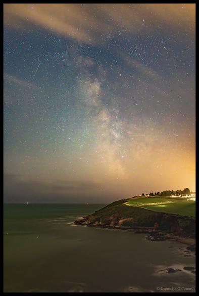 Astrophotography image showing the Milky Way galaxy stretching across a star-filled night sky above the rocky coastline and beach of Church Bay, County Cork, with meteor streaks visible and coastal lights in the distance.