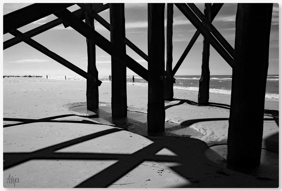 Schwarzweiße Aufnahme unter einem Pfahlbau am Strand von St. Peter Ording. Zu sehen sind die vertikalen Pfähle und einige Querstreben. Die Konstruktion aus Holz wirft starke Schatten, das Ganze wirkt sehr geometrisch. Im Hintergrund die Küstenlinie des Meeres und einige Silhouetten von Menschen.

Black-and-white photograph taken beneath a pile dwelling on the beach at St. Peter Ording. The vertical piles and several crossbars are visible. The wooden structure casts strong shadows, giving the whole scene a very geometric appearance. In the background, the coastline and several silhouettes of people can be seen.