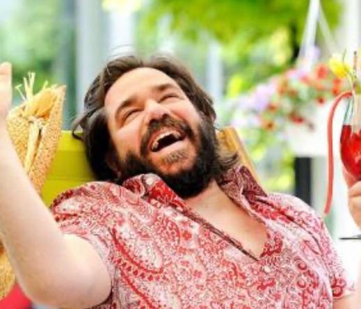A picture of Matt Berry smiling with a bright red and white shirt. He has a beard. He’s holding a red drink.