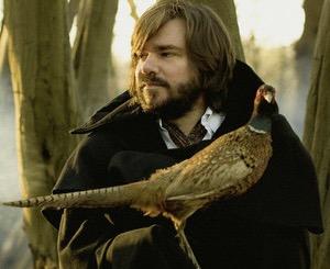 A photo of Matt Berry holding a bird in a misty forest. He has a black overcoat.