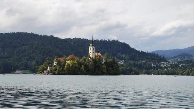 Isla del lago Bled, una pequeña isla en medio de un lago natural que aloja una iglesia. En la foto se ve el lago, en el centro de la foto la isla con la espigada torre de la iglesia sobresaliendo de la vegetación y, al fondo, montañas cubiertas de masas forestales, todo verde. En el cielo luce el sol aunque se ven nubes de tormenta (de hecho, al día siguiente cayó un aguacero importante, con acompañamiento de rayos y truenos)
