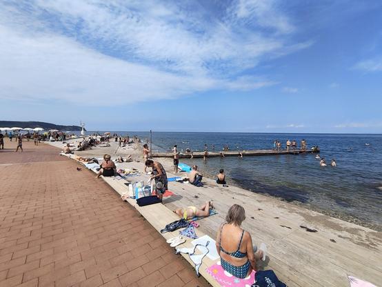 Vista panorámica de la playa de Piran, a un lado de la foto se puede ver el paseo marítimo y el resto es una playa estrecha y alargada de guijarros con un espigón que se adentra en el mar, para uso y disfrute de los bañistas. El agua es clara y esmeralda, y el cielo está azul con unas nubes finas que apenas bloquean el paso de los rayos solares. Hay una ocupación media, gente distribuida en la zona de manera cómoda y respetuosa. El agua está bastante templada y con alto contenido en sal (comprobado personalmente ;)