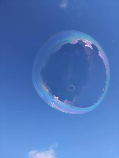 A high-angle close-up shot of a single, iridescent soap bubble floating against a clear blue sky. The bubble is nearly perfectly spherical and transparent, with vibrant rainbow colours shimmering on its surface. The colours shift and change across the bubble's film. A few small, wispy white clouds are visible in the distance, both in the sky itself and reflected within the bubble's surface. A faint, dark reflection of a tree and a house can also be seen in the bubble.