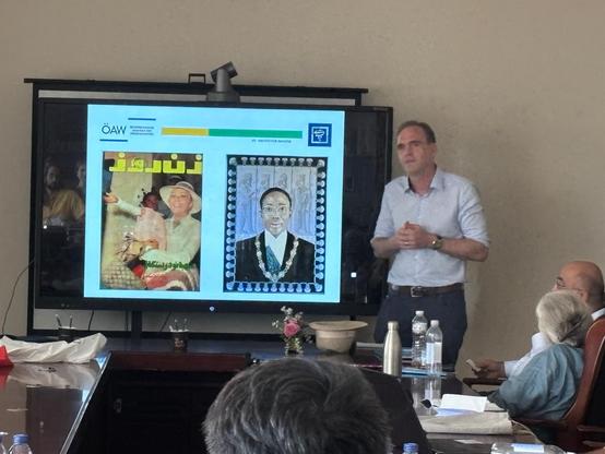 Dr. Robert Steele's presentation at the conference: He is standing just right of the centre of the picture, with hands clasped together and a powerpoint visible immediately to the left showing some late 20th century Iranian covers or book images. Several other researchers can be partially seen in the foreground in the audience.