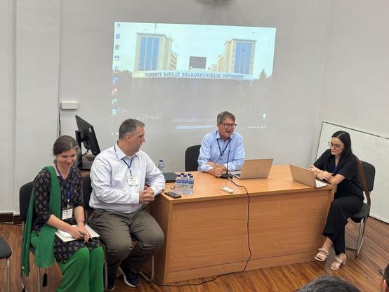 Professor Florian Schwarz leads a panel at the conference. He is sitting in the centre of the picture behind a desk, with a powerpoint projected on the the wall behind him: three other researchers are sitting around the desk, one man and one woman to the left and a woman on the right.