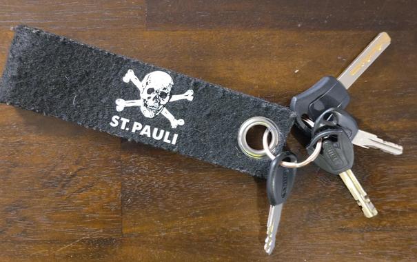 Schlüsselbund mit vier Fahrradschlüsseln und einer schwarzen Filzschlaufe mit weißem Aufdruck: Totenkopf und gekreuzte Knochen und Schrift St. Pauli.