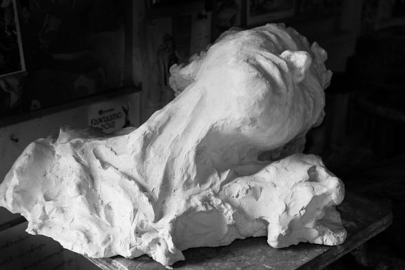 A black-and-white photograph of a plaster bust of a woman. The woman lies with her head tilted to the right, backward. The lower edge of the sculpture is roughly rendered and extends to her shoulders. The light comes from the left and falls on her turned neck and chin. The background is black. It creates a dramatic image, as if the woman is suffering.