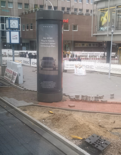 eine Baustelle neben der Fahrbahn. im vorderen Bereich ein Bereich mit offenem Sandbett. danach ein Radweg, der aus roten Pflastersteinen bestehen und auf der Linken Seite noch nicht fertig verlegt ist. dahinter eine große Fläche mit grauen Pflastersteinen. Im noch nicht fertig gepflasterten Fahrradweg steht zu 2/3 auf der Radwegfläche eine Werbesäule, die noch Recht neu aussieht und den Radweg mutmaßlich an der Stelle unbrauchbar macht.