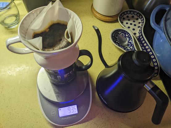A pour over coffee, into a microcenter store mug. The coffee filter is smaller than needed, but making it work. In the background are polish spoon rests on the kitchen counter, with a Dutch oven and utensil crock just inside the frame