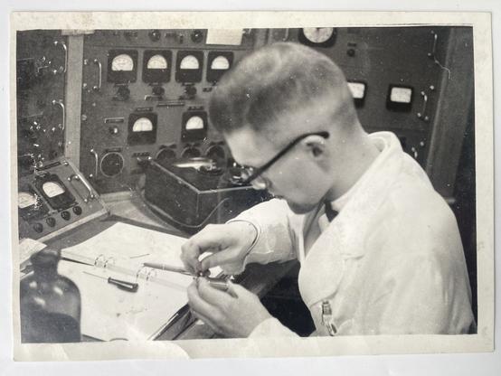 A man with black rimmed glasses and white lab coat sits at a table with some device in his hands, maybe a coil. On the table in front of him an open folder, maybe some technical manual. Behind him are many black framed analog instruments on metal panels. He is looking focused at his work.