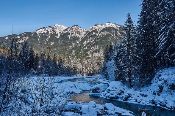 Looking over the snow-wrapped Nicolum River towards Jorgenson Peak. Filtered sunlight is reflecting on the river. The evergreen hillside and mountain are in full sun, under a clear blue sky.