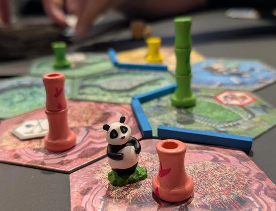 A board game panda token on a colourful field of pink, green and yellow tiles with similarly coloured bambu shoots.