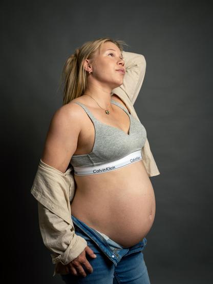 Pregnant woman with long blonde hair in a side profile view, wearing a gray sports bra, unbuttoned jeans, and an open beige shirt. She holds her hair with one hand and looks upwards. The background is plain gray.