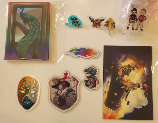 A photograph of merchandise arrayed on a white background. Pictured are: a notecard front depicting a peacock standing on a branch emerging from a picture frame, surrounded by a rainbow background; an acrylic pin in shades of blue depicting a wave; two acrylic pins with cute dragons, one in the colors of the Gilbert Baker Pride Flag, one in the colors of the Intersex-Inclusive pride flag; a standee of two chibi white men in hockey shirts holding hands; a magnet of a feather colored in shades of silver and rainbow; a sticker of a mask, one side in sunshine colors, the other in moonlight colors; a magnet of a musketeer's torso shown before a coat of arms, their sword impaling three hearts; a magnet of a dragon in the colors of the polyamory pride flag, holographic glimmers overlaying it, and a postcard of two people tumbling from a stone rook-like tower that's aflame.