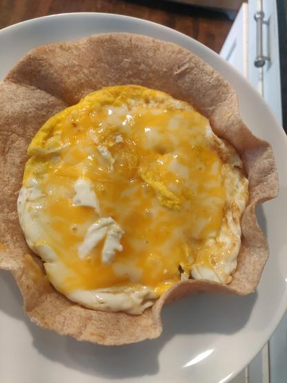 Top view, showing a fried egg with a slice of melted cheese on it, on a tortilla shell. Half of the tortilla shell has upturned edges, the other half is partially upturned.