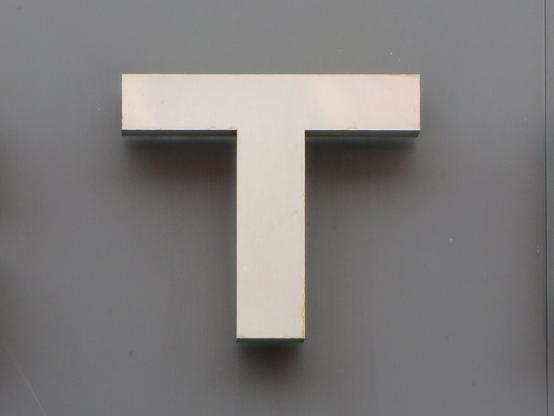 Металлическая буква Т без засечек на серой стене. Между буквой и стеной виден зазор. // A metal sans-serif T on a gray wall. A gap is visible between the letter and the wall.