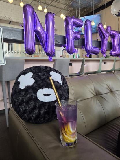 A black stuffed ball with white X-shaped eyes and a round mouth sits on a couch, pretending to drink a purple & lemon drink in a tall glass with a bronze straw. Behind the pillow are blown up purple balloon letters