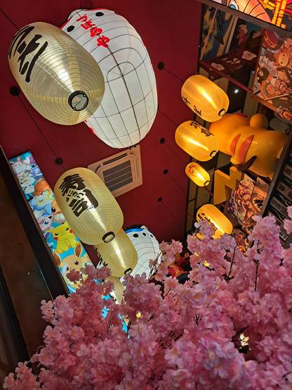 [pt] Teto de um restaurante com diferentes lanternas japonesas, bonecos de desenhos animados japoneses e uma planta cor de rosa em primeiro plano

[en] Roof of a restaurant with different Japanese lanterns, Japanese cartoon dolls and a pink plant in the foreground