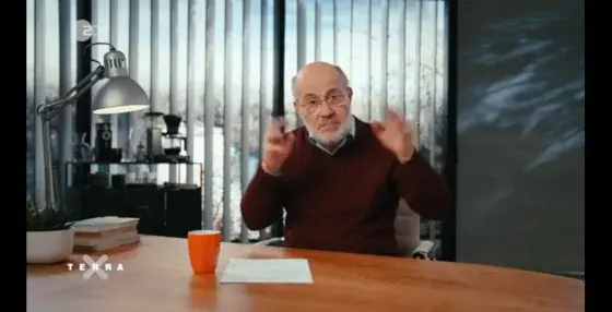 Harald Lesch sitzt an seinem Schreibtisch und erzählt. Dabei gestikuliert er mit seinen Händen. Er hat wenig Haare auf dem Kopf, aber einen weißen Bart. Und er trägt einen dunkelroten Pullover über einem hellblauen Hemd. Vor ihm steht ein oranger Becher, daneben liegt ein DIN-A-4-Papier, links im Bild sieht man eine Schreibtischlampe und einige Bücher, im Hintergrund ein kleines Regal.