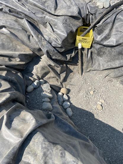 Looking down at the bottom stair. The yellow crate for the second pump is sticking into the side of the pond. on the far side. On the near side we see two lines of larger rocks arranged along the edge of the pond and bottom stair with gravel surrounding them.