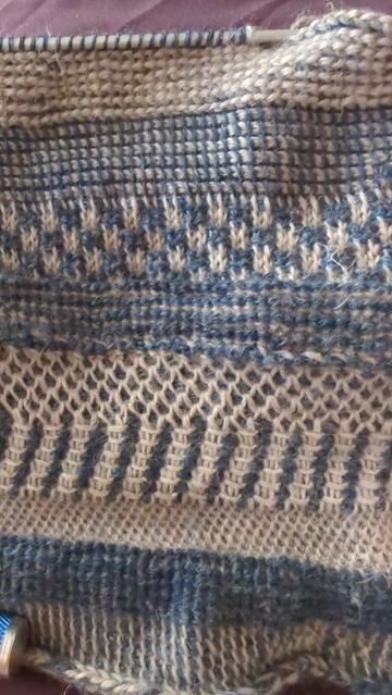 A crocheted fabric laid flat. It's made from 2 different yarns, one dark blue and one light grey. There are 9 one-inch stripes, from bottom to top, each utilizing a different stitch. Different stitches combine the 2 colors in different ways, so each band looks very different. There are ones looking like fishscale, ribbing, checkerboard, bamboo, and more.