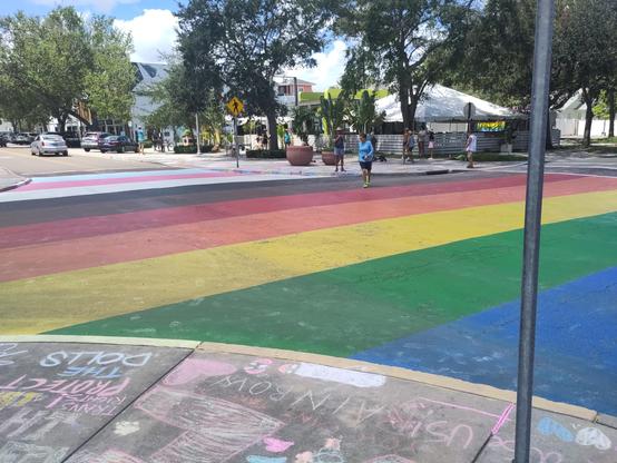 Low horizontal photo of a Pride Rainbow painted intersection surrounded by sidewalks that have hundreds of chalked images and messages of solidarity against hate.