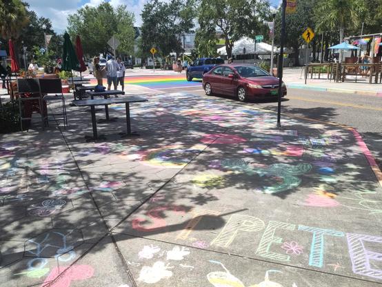 Chalked up sidewalk in the foreground is speckled with shade from live oak branches. Immediately in foreground, there is the shortened name of our city, St. Pete, in rainbow letters. Hundreds of other words and images cover the sidewalk. A burgundy sedan passes from top left to middle rich in the image, having just passed through the rainbow painted intersection in the background. People are gathered on two of the corners.