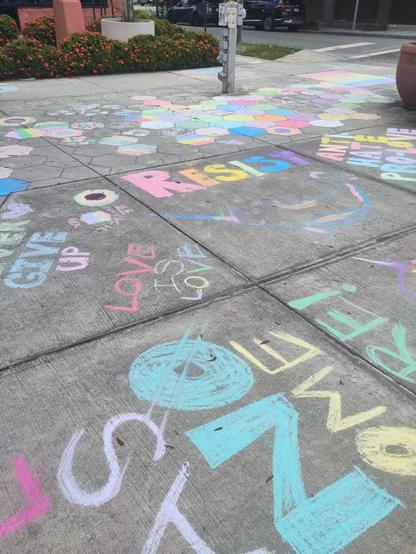 Chalked messages cover a sidewalk. In the middle, "RESIST" is written in capital rainbow letters. Some sidewalk squares are completely colored. A crosswalk is in the background, down the street three blocks from the rainbow painted intersection we were protecting today.
