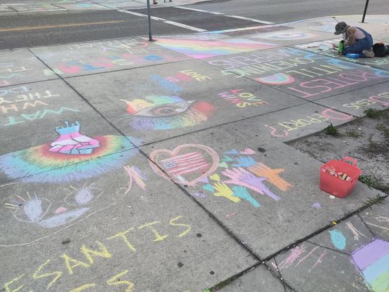 Chalked sidewalk squares in the foreground lead to a crosswalk in the background. Images on the sidewalk include a trans explicatory colored fist surrounded by a rainbow, an eye surrounded by a rainbow, a USian flag surrounded by a heart above many hands of different rainbow colors. In the foreground there is a cut off message to DeSantis. It was likely not kind.