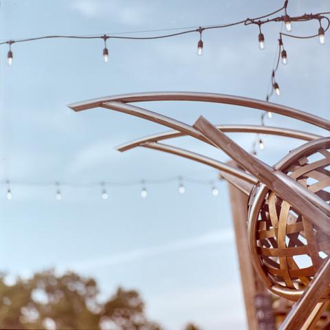 Photo of a sculpture extending from the right side of the frame towards the middle. The sculpture is made of polished steel pipes for it's framework and flat bands of bronzed metal for it's core that looks like a ball of yarn, the pipes are arching away and supported by other pipes connecting back to the core. Decorative light bulbs are strung overhead under a blue sky with stripes of clouds and very out of focus trees in the distance in the bottom left.