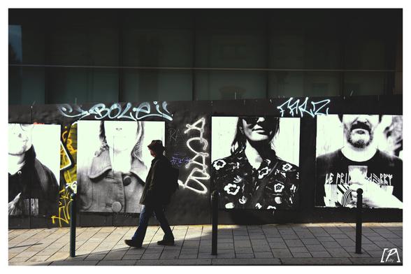 un homme marche le long du mur de la rue d el'echange, sur le mur des portraits grand format polaroids, noir et blanc, tag a c a b par dessus