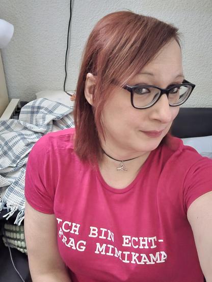 Das Bild zeigt eine Frau mit roten Haaren, die eine rote T-Shirt trägt, auf der in weißer Schrift "ICH BIN ECHT - FRAG MIMIKAMA" steht. Sie trägt eine schwarze Brille und einen schwarzen Halsreif mit einem silbernen Anhänger. Im Hintergrund ist ein Bett mit einem blau-weiß karierten Deckenbezug zu sehen. Die Frau sitzt in einem Zimmer, das mit einer weißen Wandverkleidung und einem Teppichboden ausgestattet ist.
