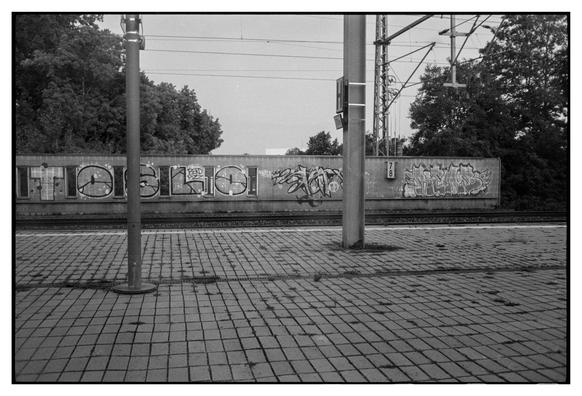 Blick aus dem Zug auf einen leeren Bahnsteig. Im Hintergrund eine mit Graffiti verziehrte Wand.