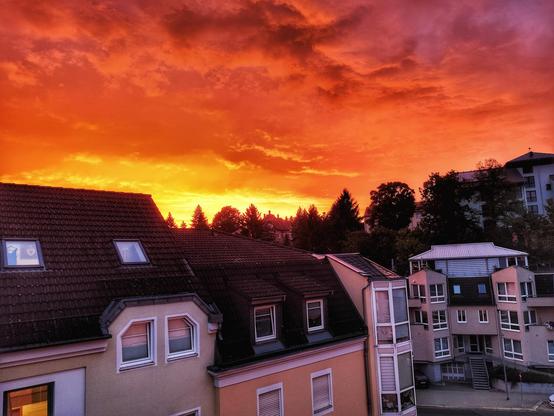 Links die Dächer von Häusern, in der Mitte hinter der Straße ein Haus, über allem der flammend rote Himmel des Sonnenuntergangs, in der Mitte am Horizont gleißend gelb das letzte Licht der Sonne.