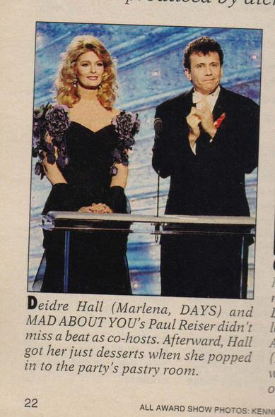 Photograph from Soap Opera Digest – April 13, 1993 showing Deidre Hall (Marlena, Days of Our Lives) and Paul Reiser (Mad About You) co-hosting an event, likely the Ninth Annual Soap Opera Digest Awards. Hall wears a black dress with floral shoulder accents; Reiser is in a black suit with a white shirt and red ribbon lapel. They stand behind a podium against a blue, starry backdrop. The caption notes their seamless hosting and Hall’s playful detour to the party’s pastry room afterward.