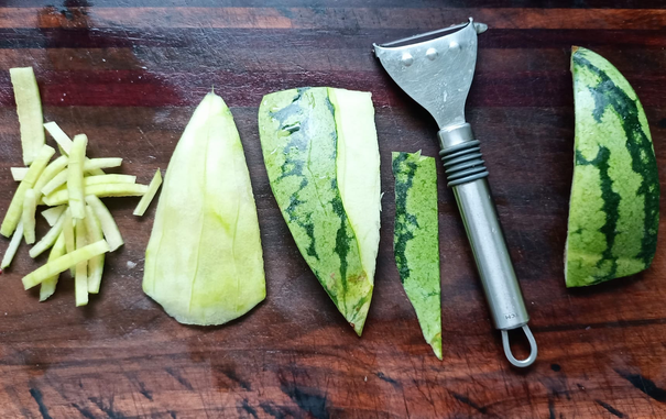 auf einem Brett von rechts nach links:
ein Stück Melonenschale
ein Sparschäler
Ein Stück Melonenschale teilweise abgeschält
Ein Stück Melonenschale abgeschält 
ein Stück weiße Melonenschale in Stifte geschnitten