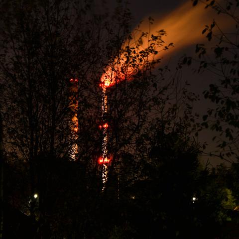 Night scene of two towering smokestacks, one of which is emitting trail of vapour, brightly visible if blurred due to long exposure. The smokestacks are obscured by dark silhouettes of trees.