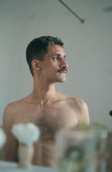 Autoportrait, torse nu devant un miroir, regardant sur le côté. En bokeh au premier plan, un blaireau à barbe et une bouture, posés sur une étagère de salle de bain.