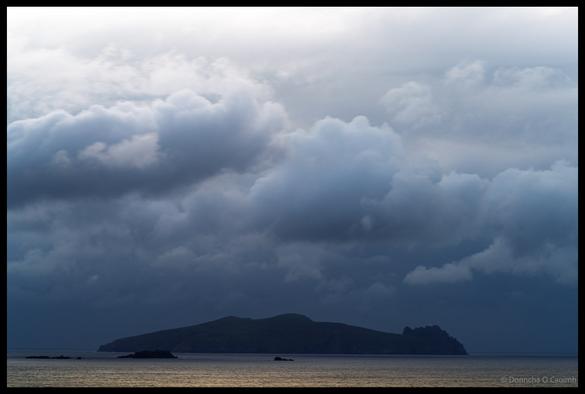 Dramatic storm clouds gathering over the silhouetted form of An Fear Marbh (The Dead Man) rock formation viewed across dark waters from the Dingle Peninsula, County Kerry.